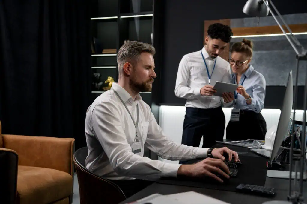 Automation manager working on a computer while two workers stand in the back and discuss reports shown on a tablet