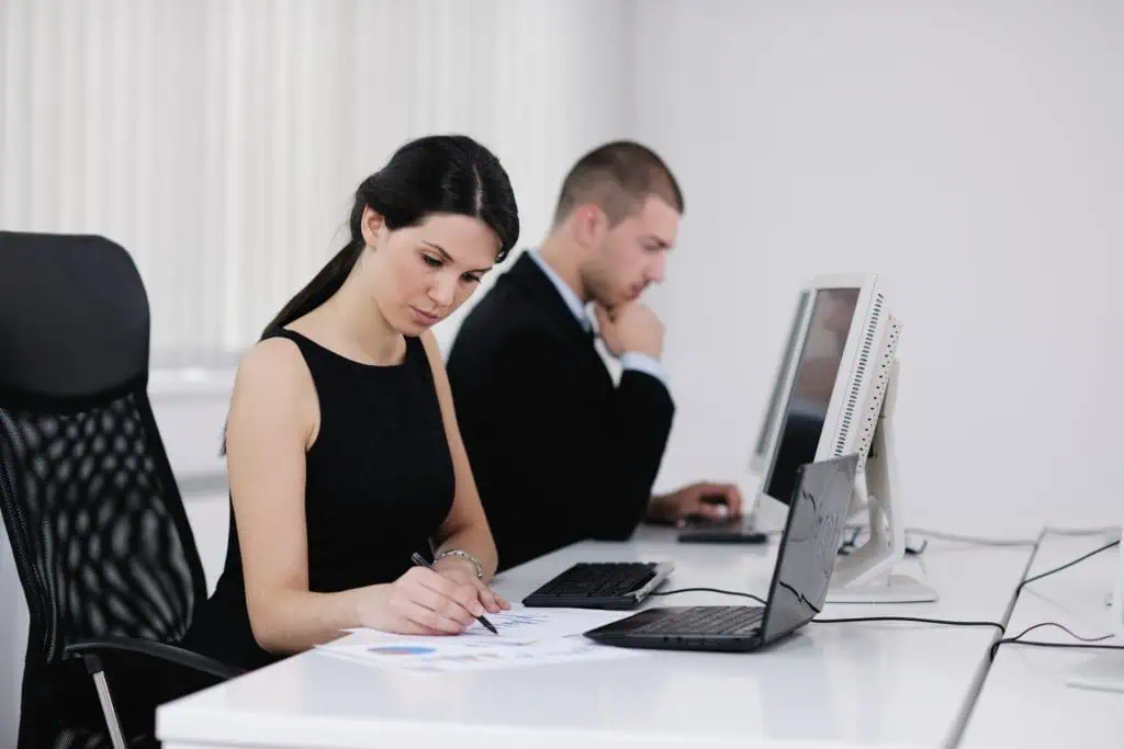 Two administrative workers, man and a woman, are sitting at a table, each working on their computer with the woman going over paper reports
