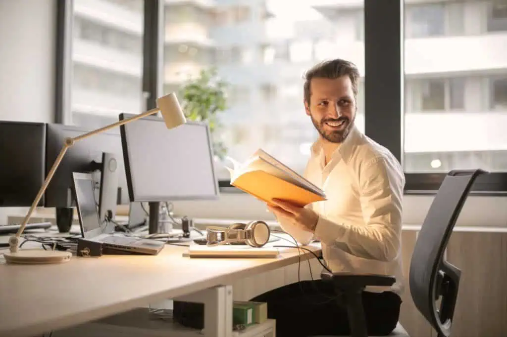 Launch manager works at his table in office, he is holding a notebook in his hand and looking over his shoulder with a smile