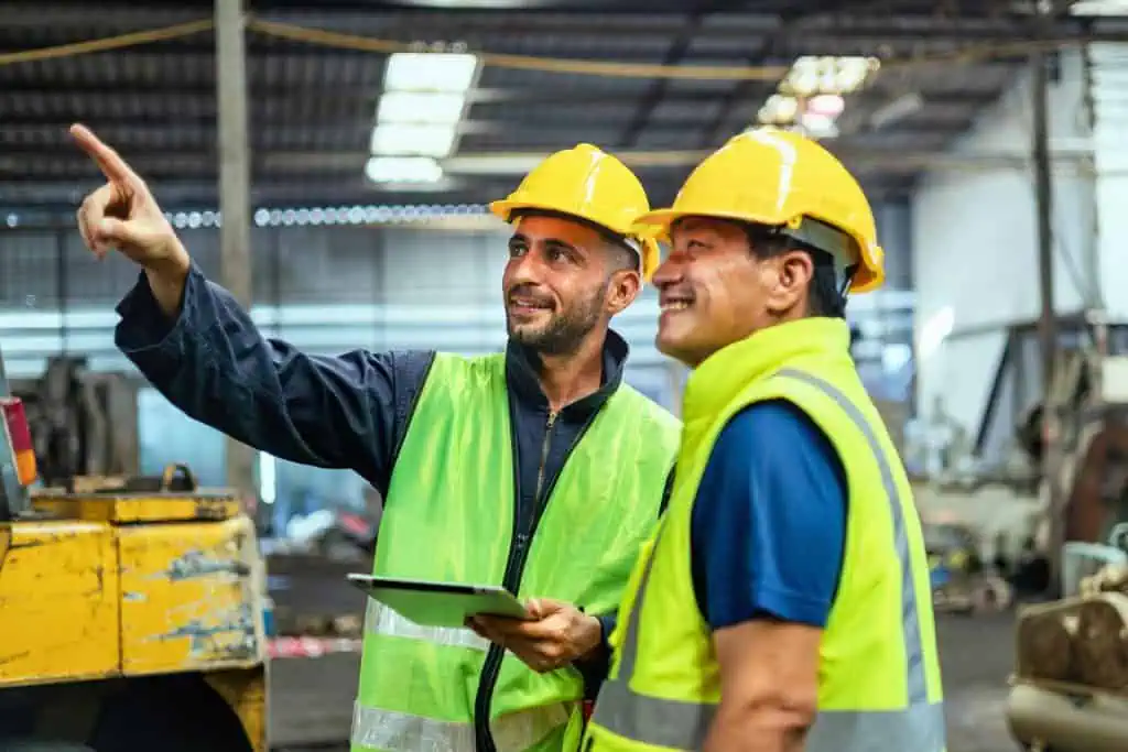 Two men in safety vests and helmets are standing in a warehouse, one holding a tablet and pointing forward