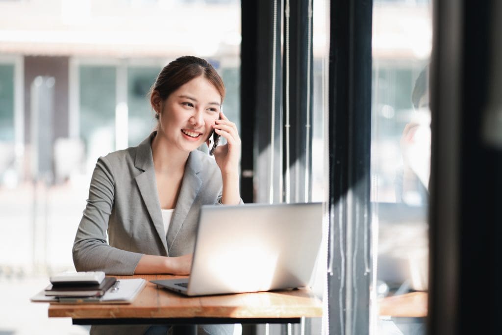 Young business woman talking on smartphone with laptop in front of her at a desk in a room with big windows.
