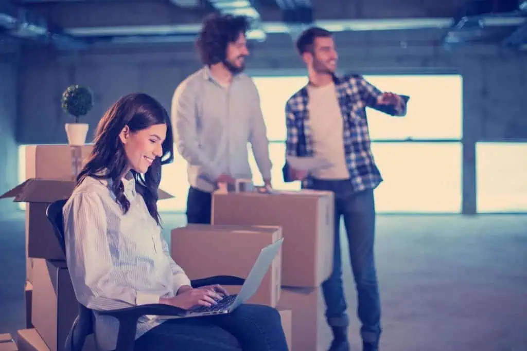 Three relocation management experts in empty building, two men stand in the backround with boxes, woman is sitting on chair with laptop in her lap