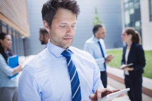 Sales executive search expert in blue shirt and tie holds tablet in his hand, scrolling. He's standing outside a building with talking business people in the backround.
